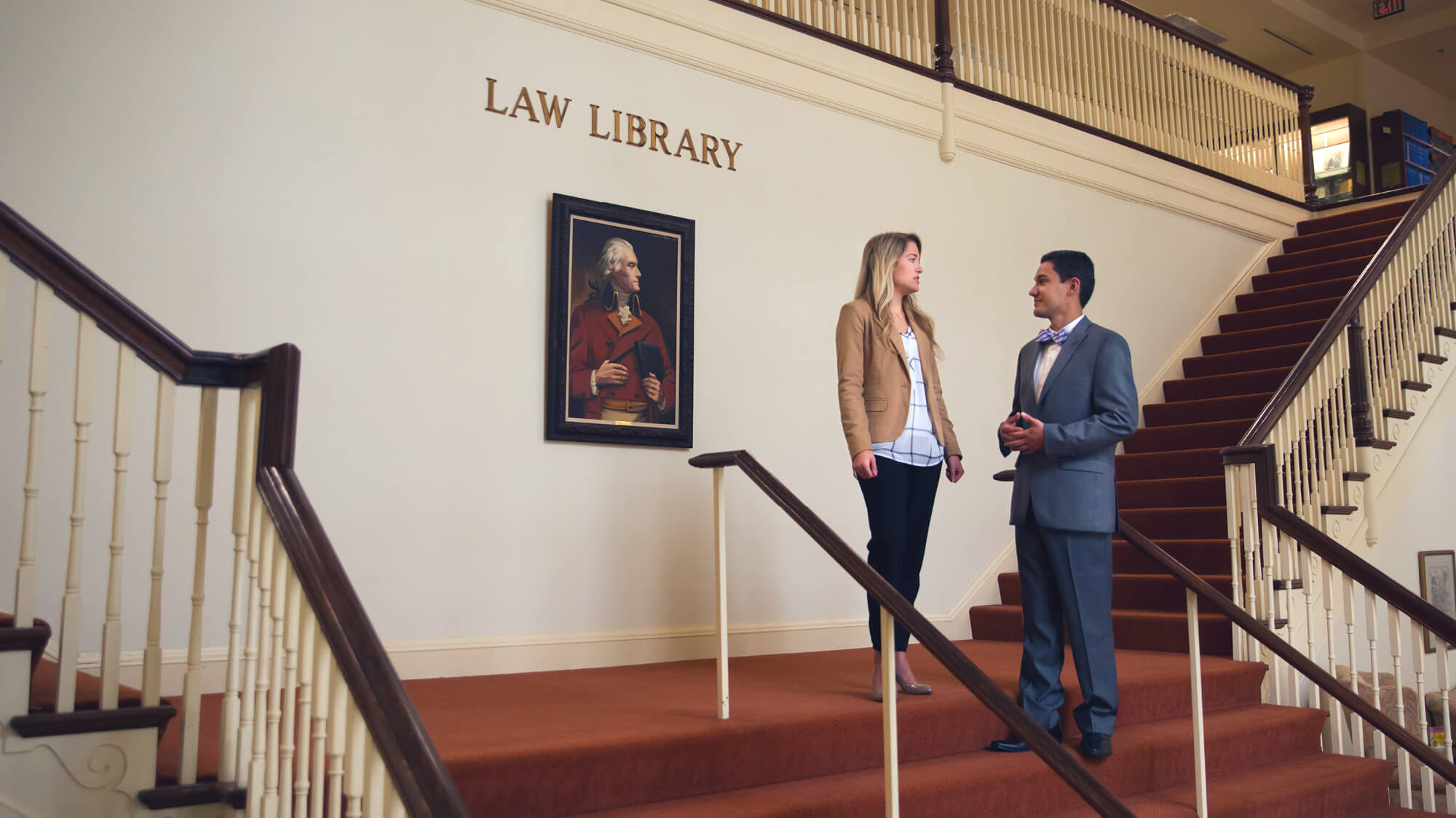 Two professionally dressed law students conversing on a staircase in the Regent University Law Library, with a painting of a historical figure on the wall behind them.